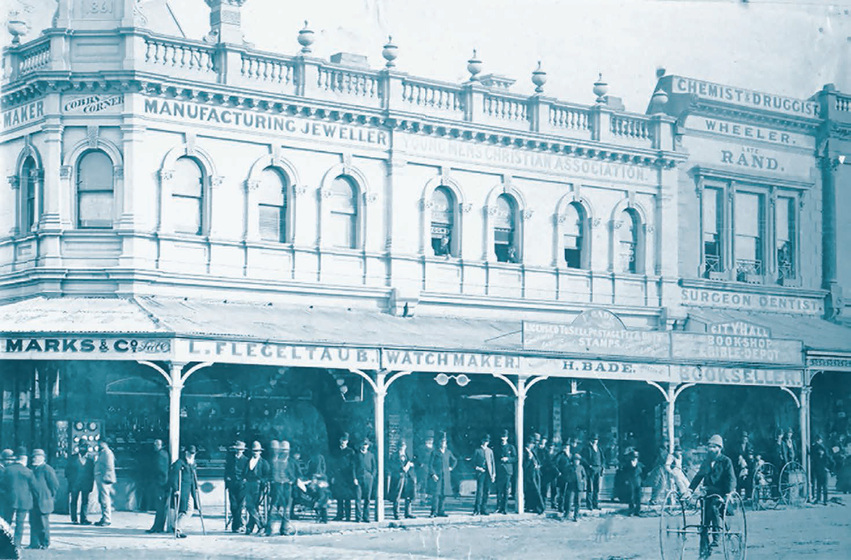 Ornate two-story building with a street in the foreground, and a crowd of people in front of the building. A man rides a bicycle in the foreground.