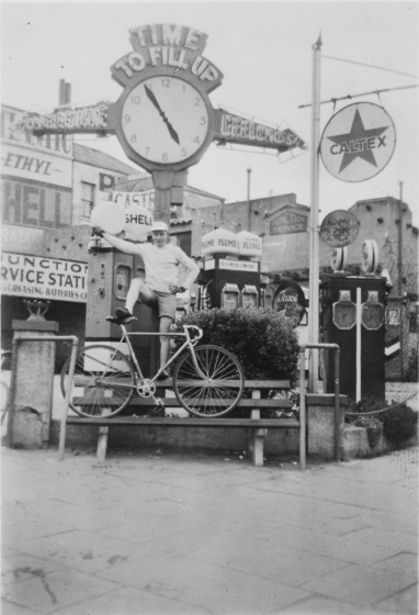 Young man stands with one foot on the seat of a bicycle, which is standing on a park bench in front of a cluster of petrol pumps or bowsers. 