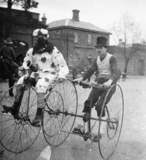 Two men riding cycling machines. The man on the left rides a penny farthing and wears a white costume with a heart pattern. The man on the right rides a three-wheeled bike with large wheels at the front, wearing a top hat and vest.