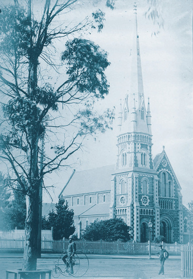 Stone church with tall spire behind a fence. In the foreground is a tall tree and a man riding a three-wheel cycling machine, with large wheels at the front.