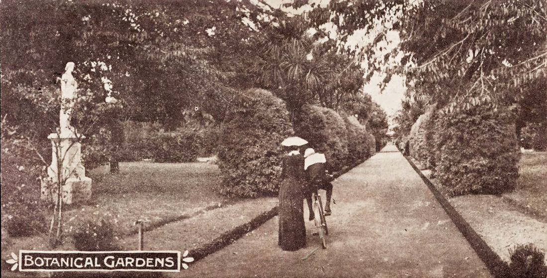Behind view of a man on a bicycle leaning on a woman in an 1890s dress, cycling down a path lined with shrubs and a statue to the left.