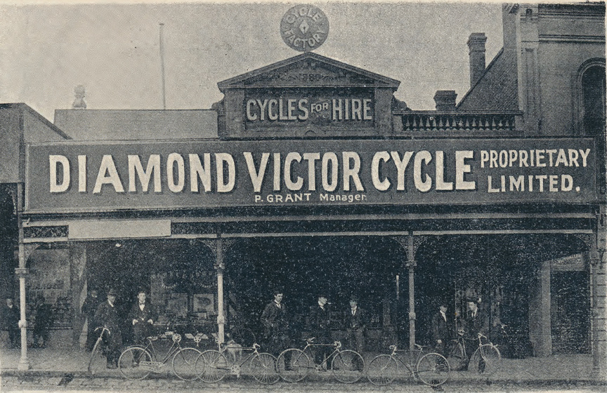 Seven men holding bicycles stand in front of a store building, with text above the roof reading 'Cycles for Hire. Diamond Victor Cycle Propriety Limited'