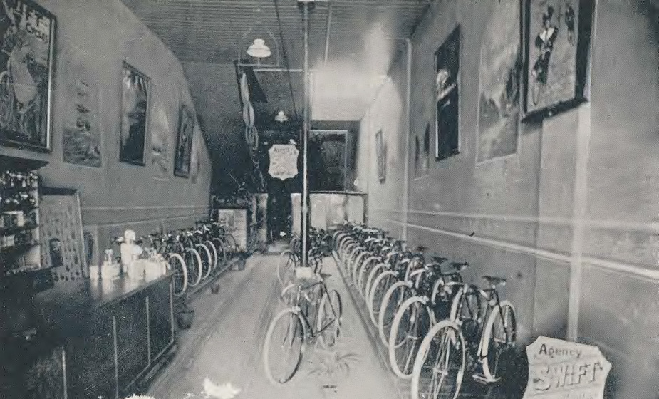 Long narrow room with rows of bicycles along each wall, a bicycle in the centre. On the walls are framed pictures. 