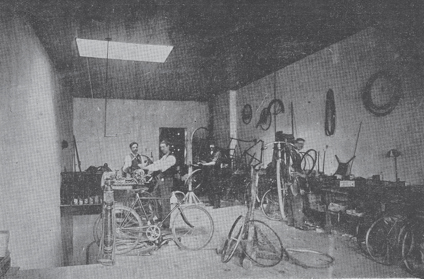 Room with many bicycles spread throughout, including lying down on the ground. Two men in long sleeved shirts, waist coats and trousers stand among the bicycles. 