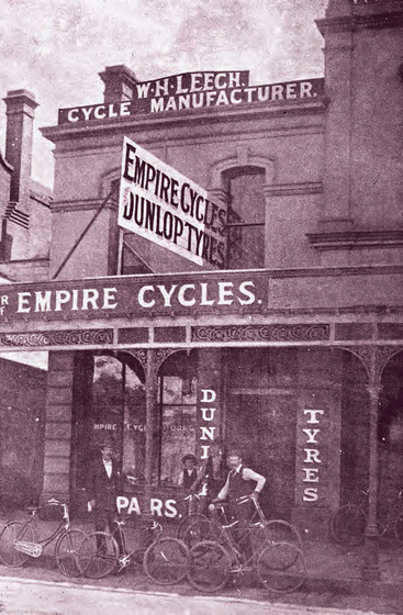 Two story building with four men holding bicycles standing in front. Building advertising sign reads Empire Cycles Dunlop Tyres'