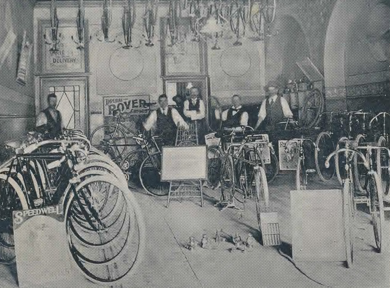 Five men stand at the back of a room with rows of bicycles and some bicycles placed throughout.