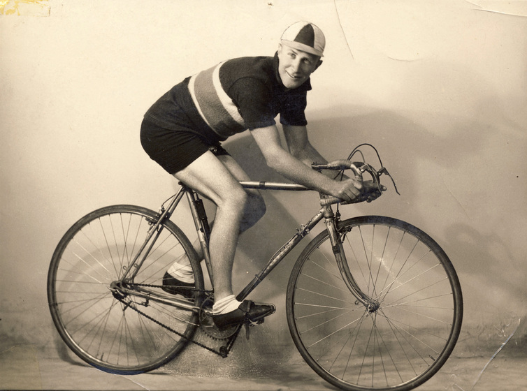 Young man riding a bicycle, wearing a racing jersey with a stripe across the chest, and a multi-panelled cycling cap.