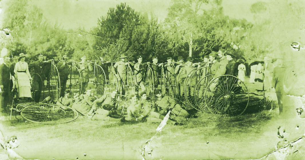 Row of men and women standing outside in 1890s outfits, holding penny farthing bicycles. Some bicycles are also laying on the ground in front of them.