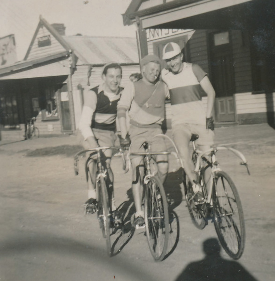 Three young men ride sitting on bicycles in a town stree with arms around each other, wearing striped racing jerseys and cycling caps. 