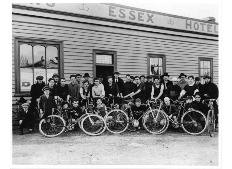 Group of men holding bicycles standing in front of a weatherboard building with a sign above reading 'Essex Hotel'
