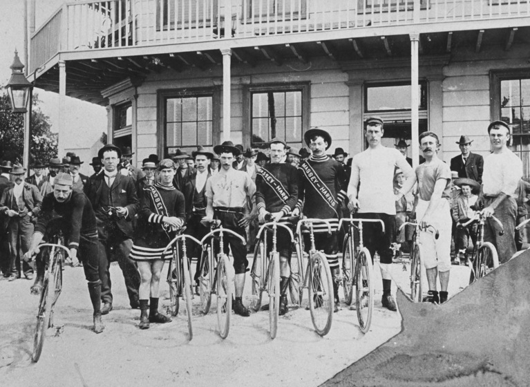 Group of men with bicycles standing in front of a two-story weatherboard building. Two men have sashes crossing their bodies.