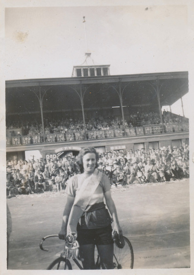 Young woman with a sash worn across her body stands with a bicycle on a sporting field with a crowd of people in the background. She stands in front of a spectator's stand.