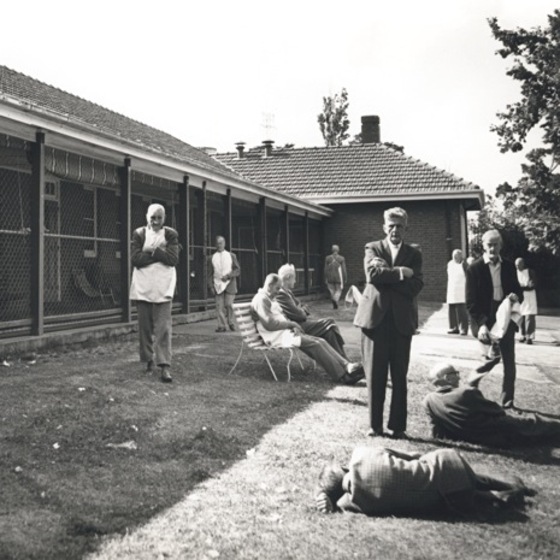 Group of men standing, sitting and laying on grass in front of a brick house with wire fence enclosed veranda. Two standing men have their arms crossed.