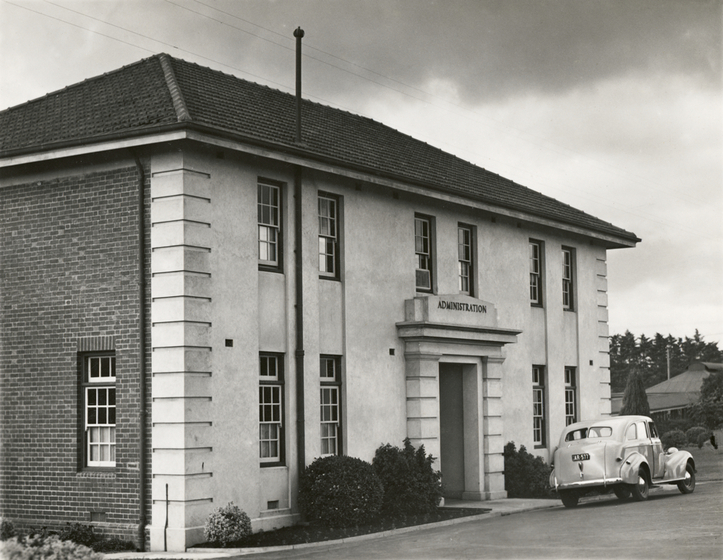 Two story brick and render building with a car parked in front and shrugs either side of the front door. The word 'administration' is above the entry door.