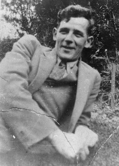 Young man wearing a suit leans back on his left elbow on grass. Trees are visible in the background.
