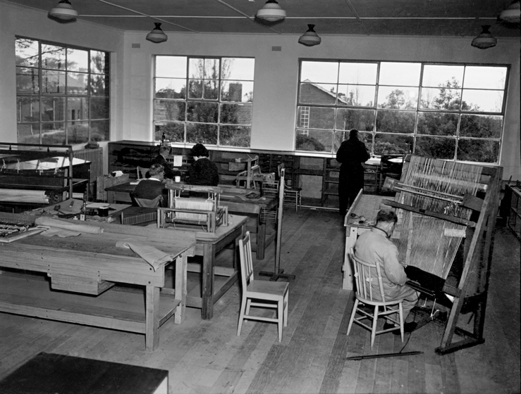 Room with wooden work bences, tables and chairs. A man on the right sits at a standing loom. Another man stands at the back of the room looking out through windows to trees and houses. Three other people sit at tables at the back.