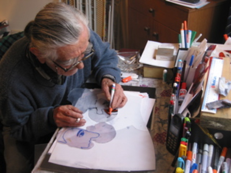 Elderly man leaning over a desk covered in markers, pens and other crafting materials. He holds a market in his hand and is drawing onto transparent film.