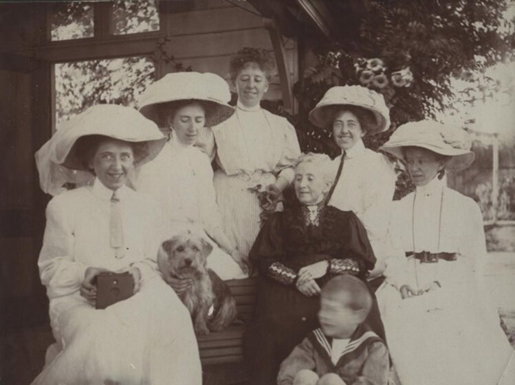 Black and white photograph of six women, one small boy and a small dog seated in front of a house. Five of the women wear white dresses, with four wearing large white hats. An elderly woman wears black sitting centre, and the boy is wearing a sailor top. 