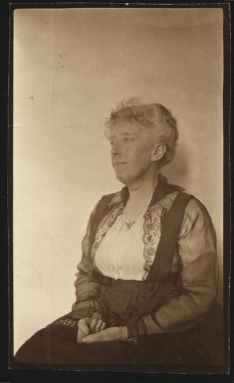 Portrait of a middle aged woman seated with hands in lap, wearing a white top with black lace over top and skirt.