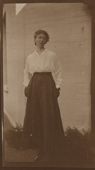 Woman with ankle length black skirt and white blouse standing in front of white brick wall.