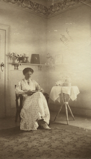 Elderly woman seated in a room with a floor rug, with table to right. She is reading a book, and the table has a table cloth, tin and flowers on it. Behind is a shelf with pictures and vases of flowers.