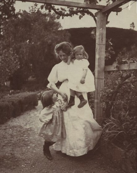 Young woman with long white dress walking in a garden with two young girls. She holds the hand of one girl to her left, and holds the second girl on her opposite hip.