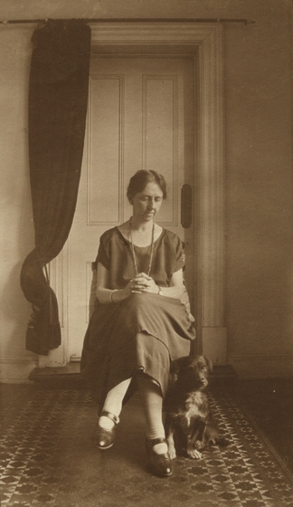 Woman in a dress wearing a necklace seated in front of a door, with a patterned rug beneath and small dog at her feet. There is a curtain to the side of the door behind.