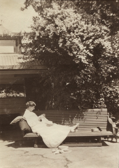 Woman in a long dress sitting with legs stretched out along a park bench. A large brimmed hat is on the ground in front of her, and a shrub and house is seen behind. 