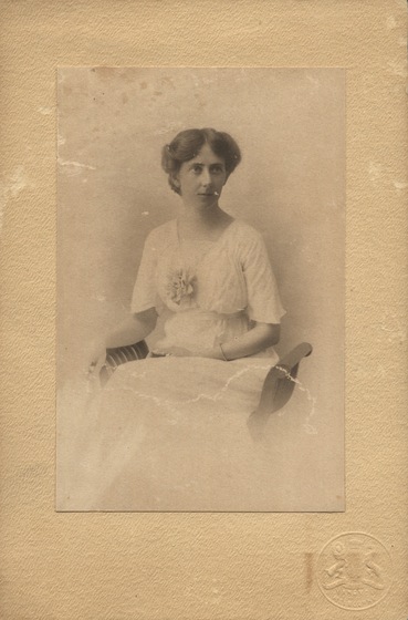 Studio portrait of a woman in a light dress, with one hand in her lap seated on an ornate chair with no back. 