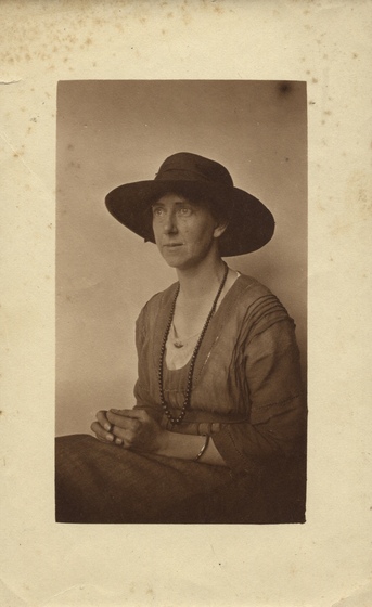 Studio portrait of a woman in a dark dress, seated with hands in her lap, wearing a large brimmed hat and beaded necklace.