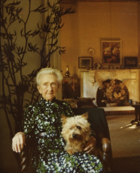 Elderly woman seated in a wooden chair wearing a patterned green, white and black dress with a small dog in her lap. Behind is seen another room featuring a fireplace with paintings above. 
