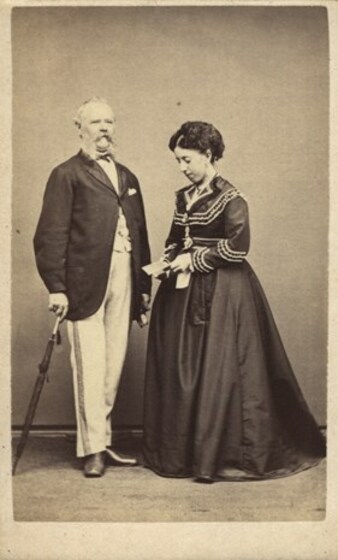 Studio portrait of a middle aged man and woman. The man wears a long coat and rests a closed umbrella to his side. The woman wears a long dark dress and holds a booklet.