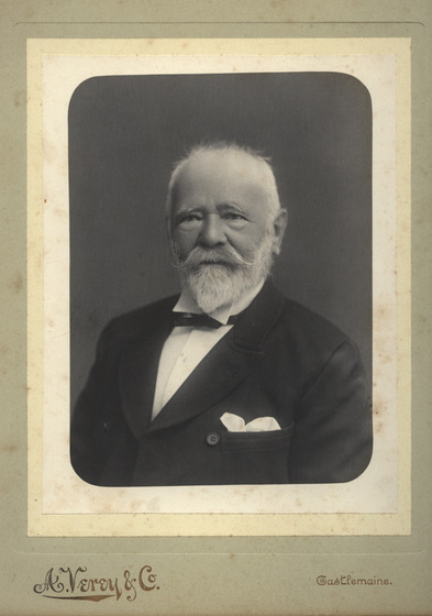 Studio torso portrait of an elderly man with a beard and moustache, wearing a dinner jacket and shirt.