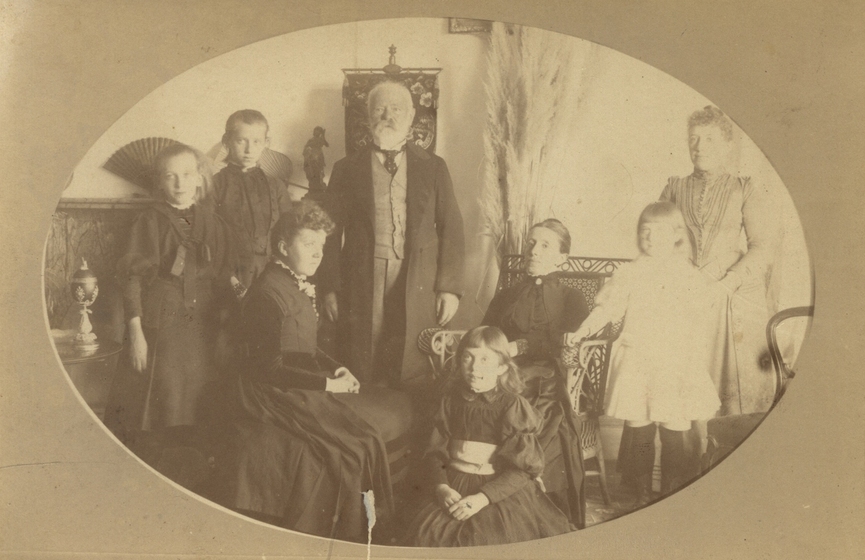 Photograph with an oval card frame, with an elderly man standing centre, surrounded by two girls to the left, a middle aged woman and child seated in front, another elderly woman seated to the right and a child and another woman standing to the right. 