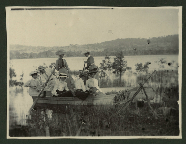 Group of six women and one man, all wearing large brimmed hats, in a row bot on a river. There is grass in the foreground, and submerged trees behind.