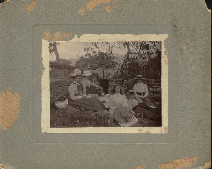 Photograph framed by card, of a group of five women and one man seated on the ground on a picnic rug. There is a basket to the right and four of the five women wear wide brimmed hats, with one holding a hat in her lap. Trees are seen behind.