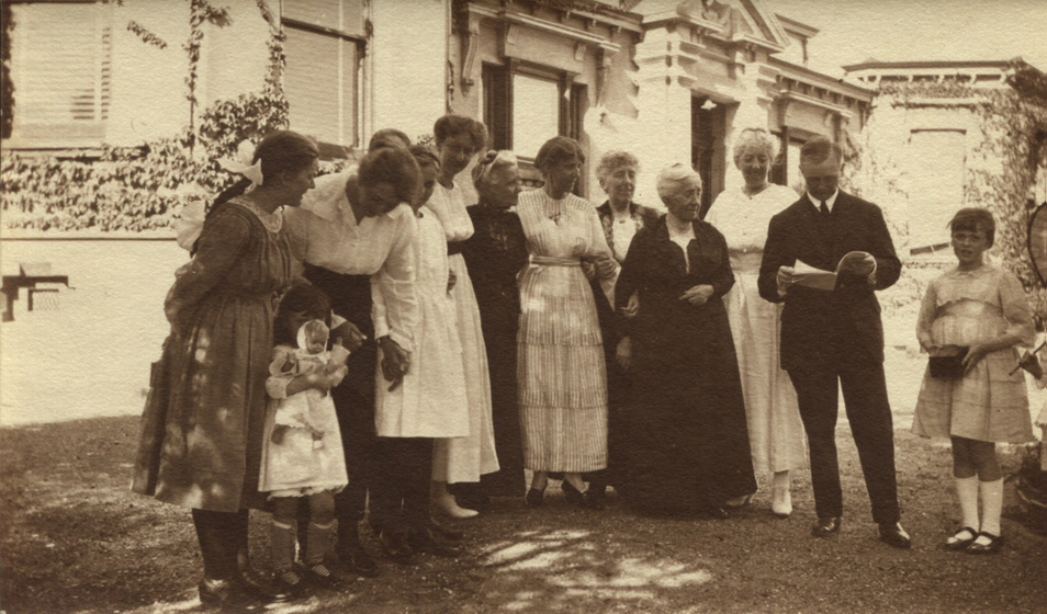 Group of nine women, two girls, and one man standing in front of a house. The man stands with a book open and reading while the women look on. One child is holding a small doll.