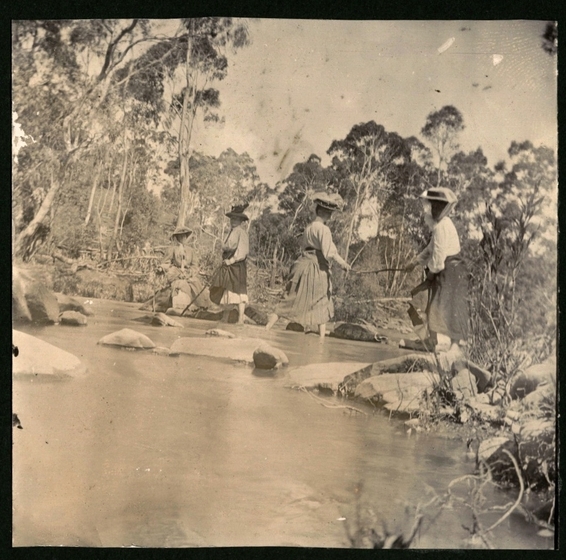 Four women in bare feet with skirts held up walking across a stream. Rocks are seen in the foreground with trees behind.