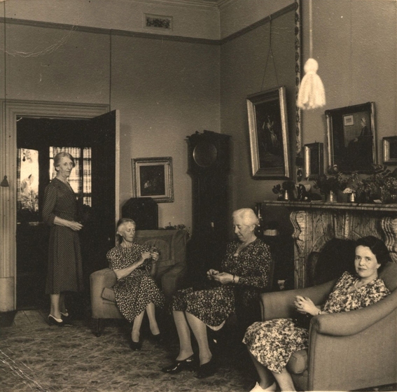 Four women in a sitting room. Three women sit in chairs, while one stands at a door on the left. Behind are paintings, a grandfather clock, and a fireplace.