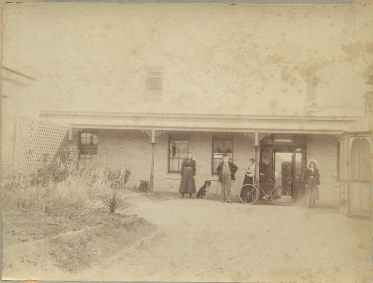 Small brick house with chimney with three women and a man standing in front, with a dog and bicycle. A garden bed is seen in the foreground.
