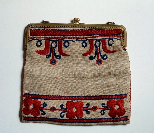 Small aged cream fabric bag with metal clasp at top, with three embroidered red flowers with blue trim along the bottom, and a pattern of red and blue embroidery along the top. 