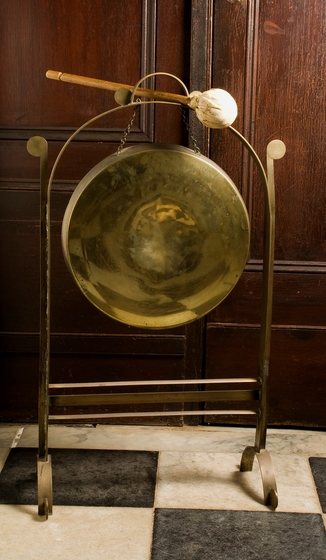 Metal gong on a stand, standing on a black and white check floor with wooden panelling behind and a stick with a padded end resting on top.