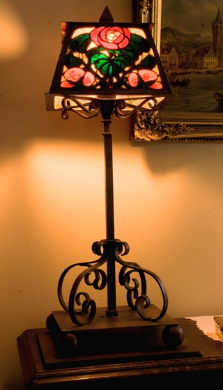 Lamp with light turned on, with ornate curled base and a lead-light shade featuring red roses with green leaves.