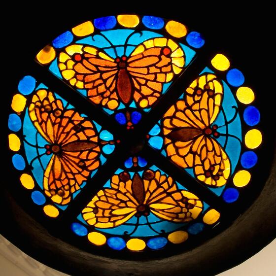 Circular stained glass pattern, featuring four mirrored orange butterflies on blue backgrounds. Yellow and blue circles surround the design. 