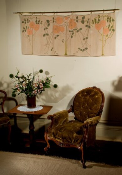 Fabric with pattern of embroidered red flowers and green leaves and vines hanging from a metal rod on a wall. In front is an ornate upholstered moss green coloured chair and side table with a vase of flowers on it. 