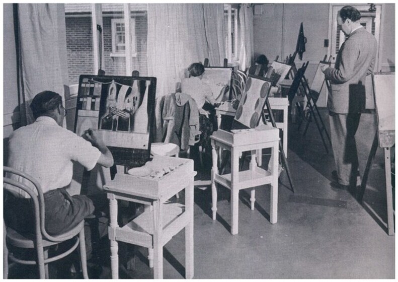Three patients painting on art easels. To the right of the artists is a small bench with a set of paints to use. Nearby a Medical Professional supervises.