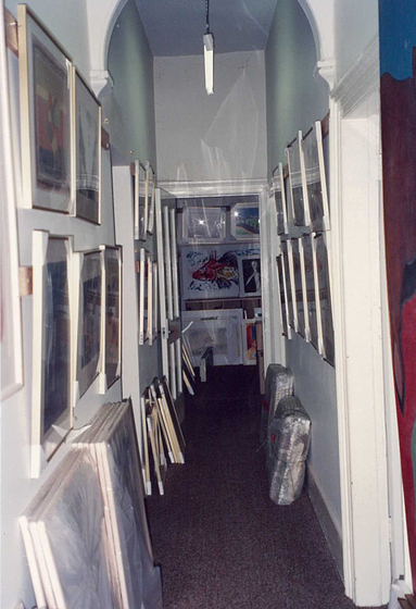 Hallway of Faraday Street terrace, showing wrapped, framed works on paper stacked up against each wall. More framed works on paper hang in two rows along all walls of the hallway.