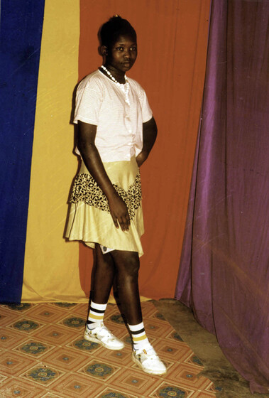 Young woman stands on a brightly coloured tile floor, with blue, yellow, red and pink cloth hanging behind. She wears a short yellow satin skirt, white blouse, and white bead necklace.