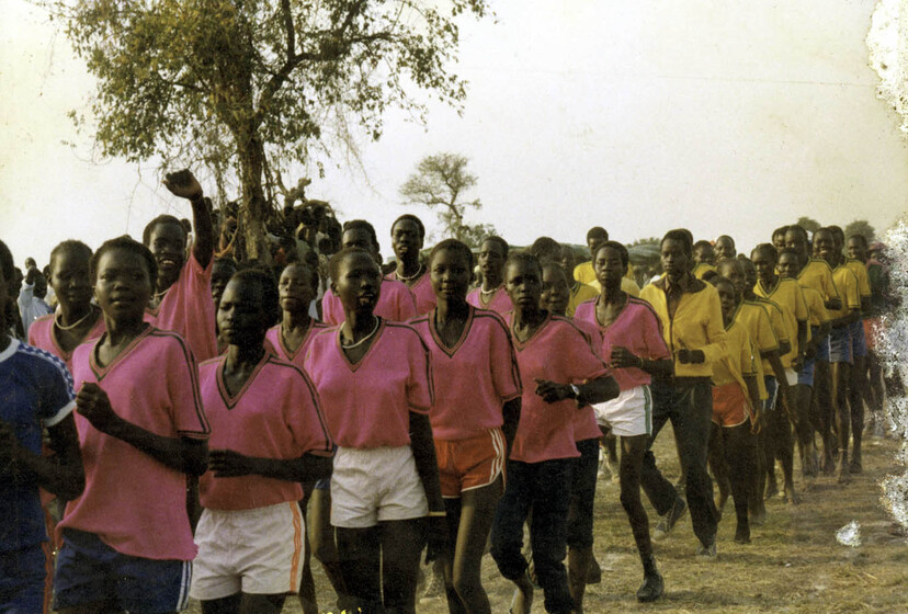 Two lines of young women, running with a tree behind in the background. The front group of women wear a uniform of pink shirts and white shorts. The back group of women wear uniforms of yellow jackets and blue shorts.