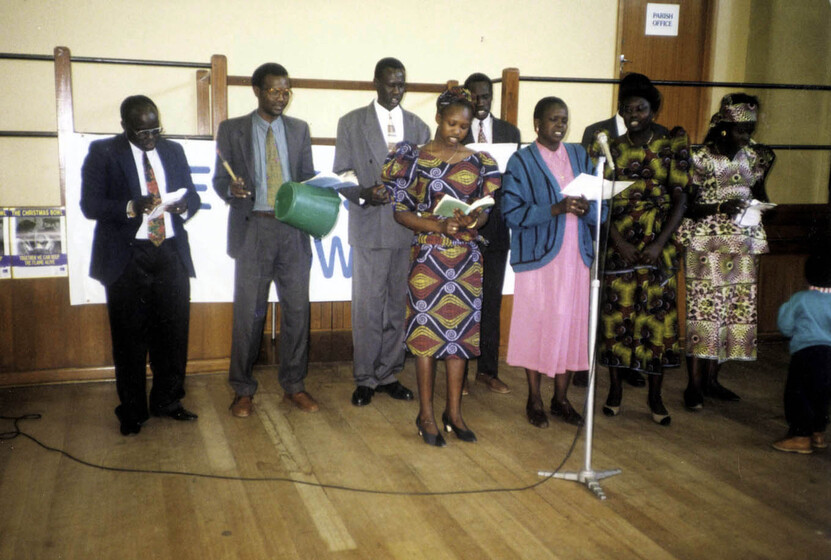Group of men and women standing in a hall with timber floors behind a single microphone and stand. Four men wear suits, and one carries a bucket upside down with a stick. Four women wear brightly patterned dresses and three hold books open.